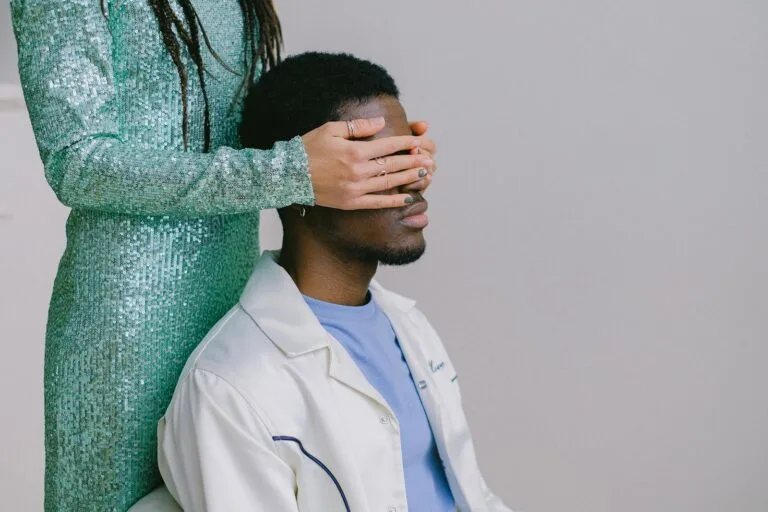 A woman in a sparkling green dress surprises a man by covering his eyes indoors.